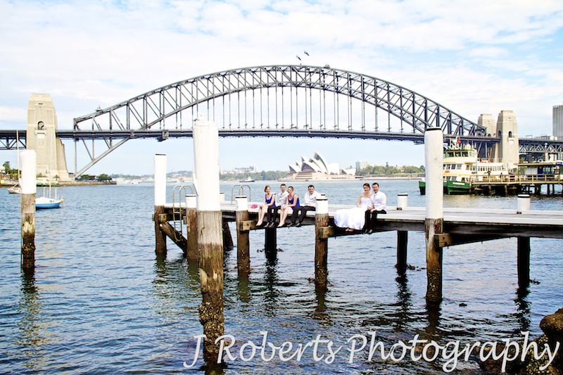 Bridal party sitting on edge of pier with Sydney Harbour Bridge, Opera House and ferry in the background - wedding photography sydney